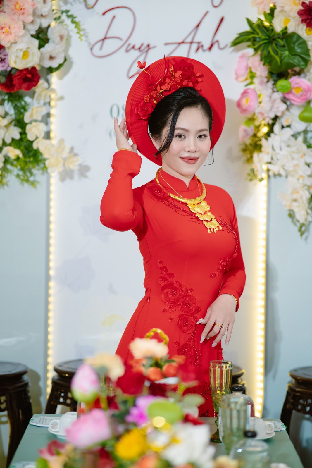 Cô dâu áo dài đỏ rạng rỡ bên backdrop hoa trong lễ gia tiên
