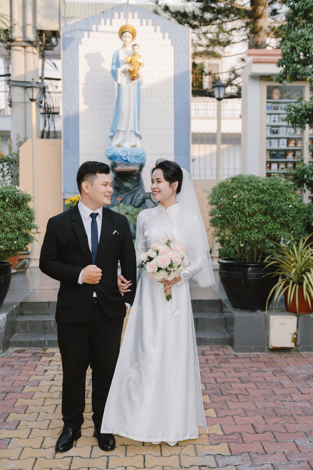 Cô dâu áo dài trắng và chú rể đứng trang nghiêm trong sân nhà thờ trước lễ Hôn Phối