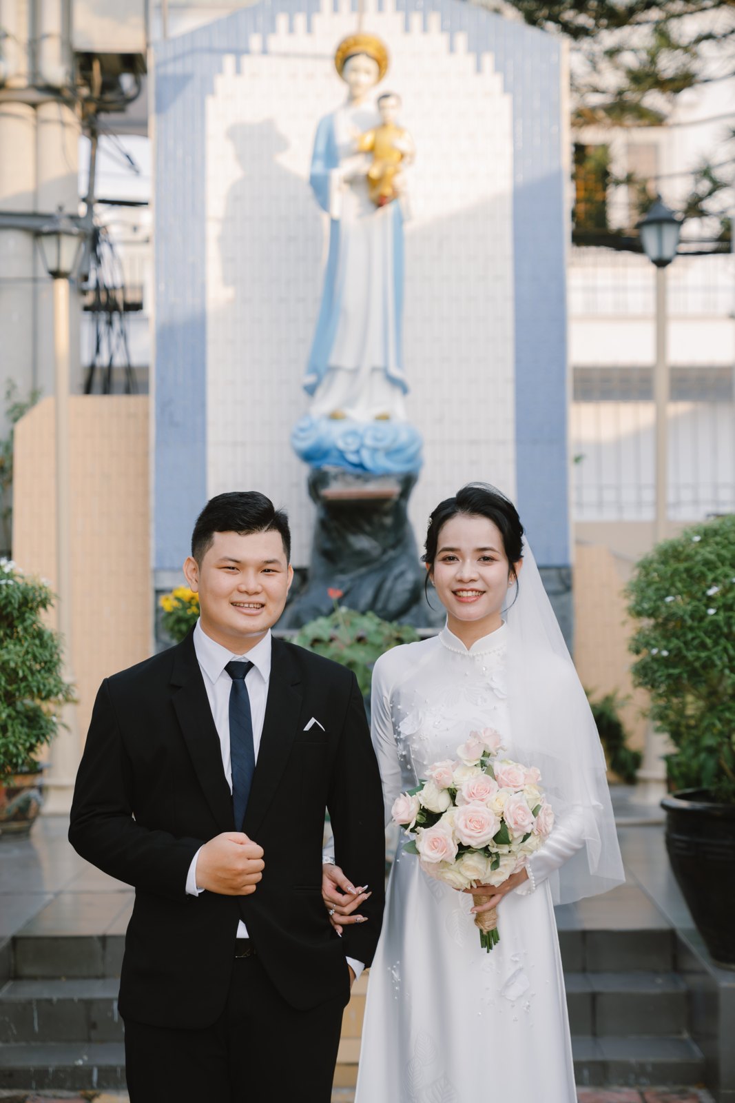 Cô dâu chú rể Công giáo bên tượng Đức Mẹ trong khuôn viên nhà thờ ngày lễ cưới